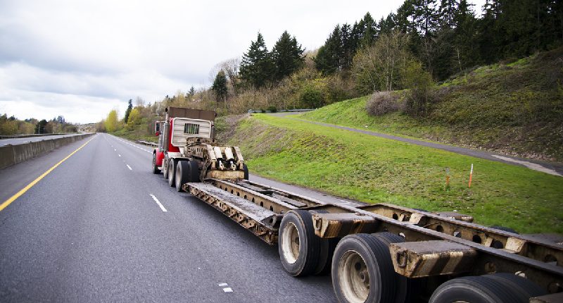 Red semi-truck hauling a large, empty flatbed trailer on a rural highway with green trees and grass-covered hills in the background.