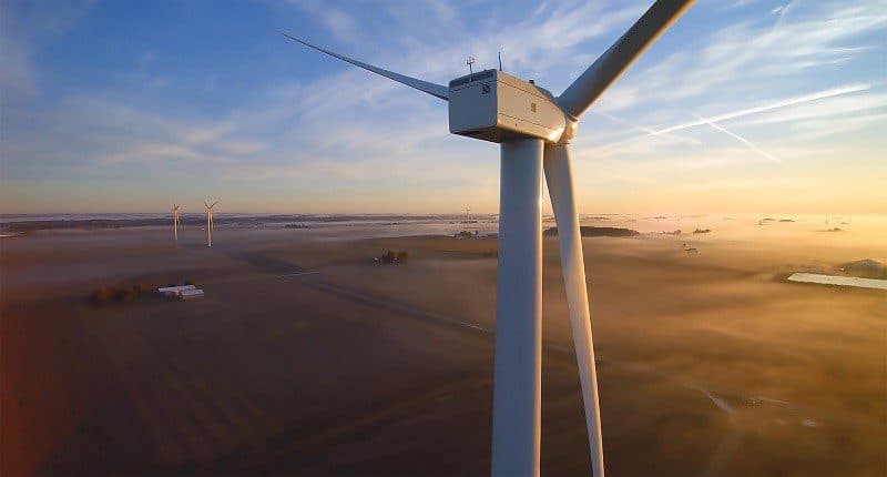 Aerial view of a wind turbine in a vast rural landscape during sunrise, with clear skies and other turbines visible in the distance.