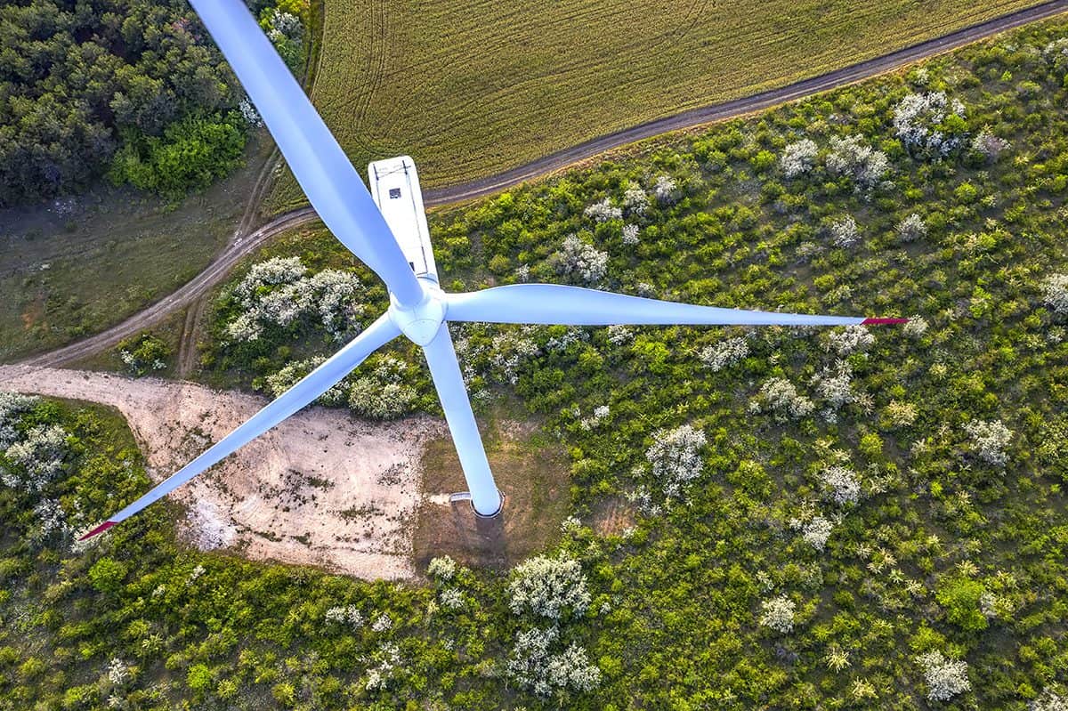 Aerial view of a wind turbine standing over lush green landscape with pathways and trees visible below.