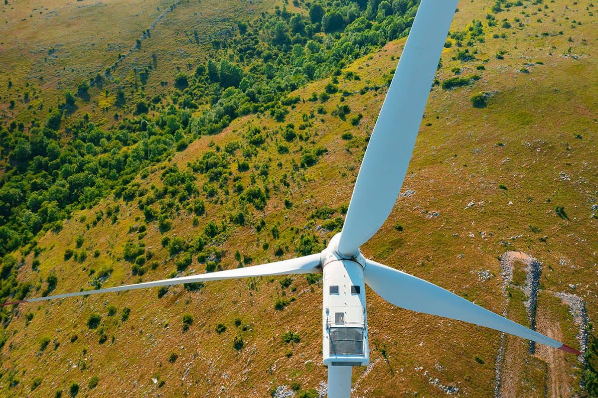 Wind turbine with large propeller generates electricity Aerial view of a wind turbine standing over lush green landscape with pathways and trees visible below.