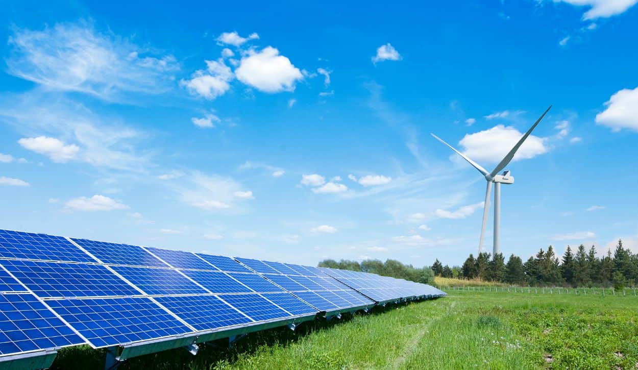 Row of solar panels in a sunny field with a large wind turbine in the distance