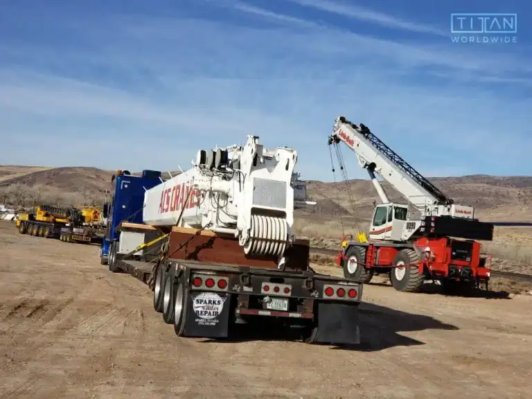 Flatbed truck transporting heavy crane equipment in a rural landscape with mountains and additional construction vehicles in the background.