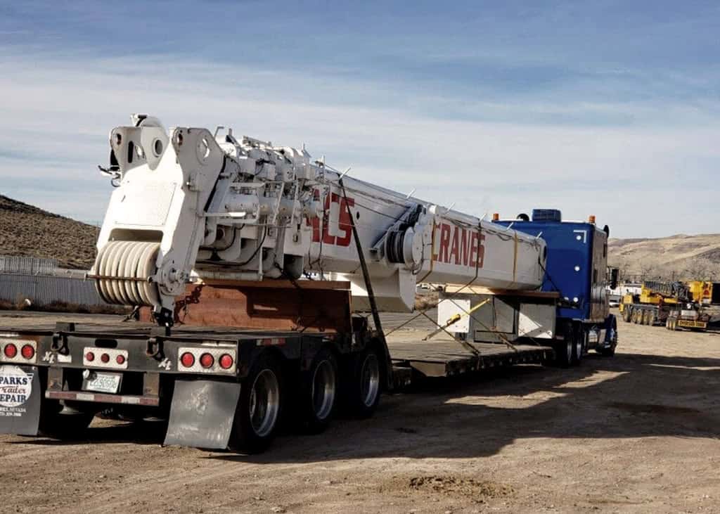 Large crane mounted on a truck trailer, parked on a dirt road in an industrial area with hills in the background.