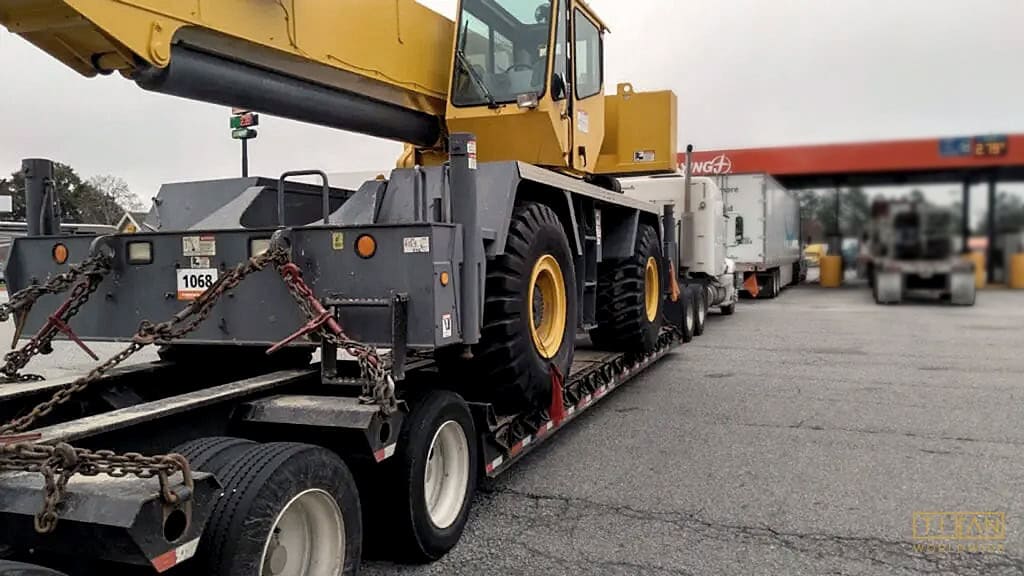 Yellow crane on a flatbed trailer at a fuel station, secured with chains, behind a truck.