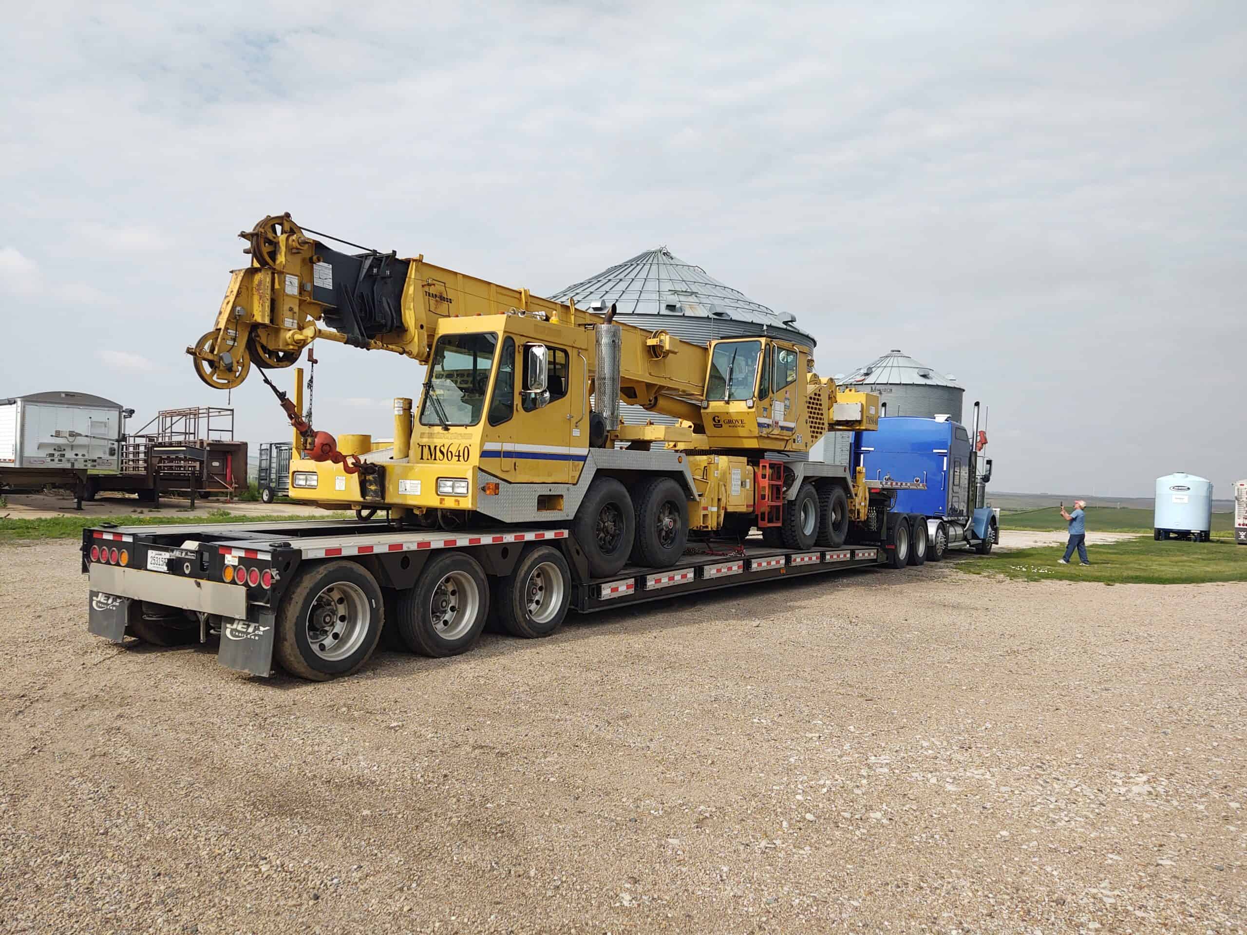 A Grove TMS640 hydraulic truck crane loaded on a lowboy RGN trailer, secured for transport at a rural job site with grain silos and farm structures in the background.