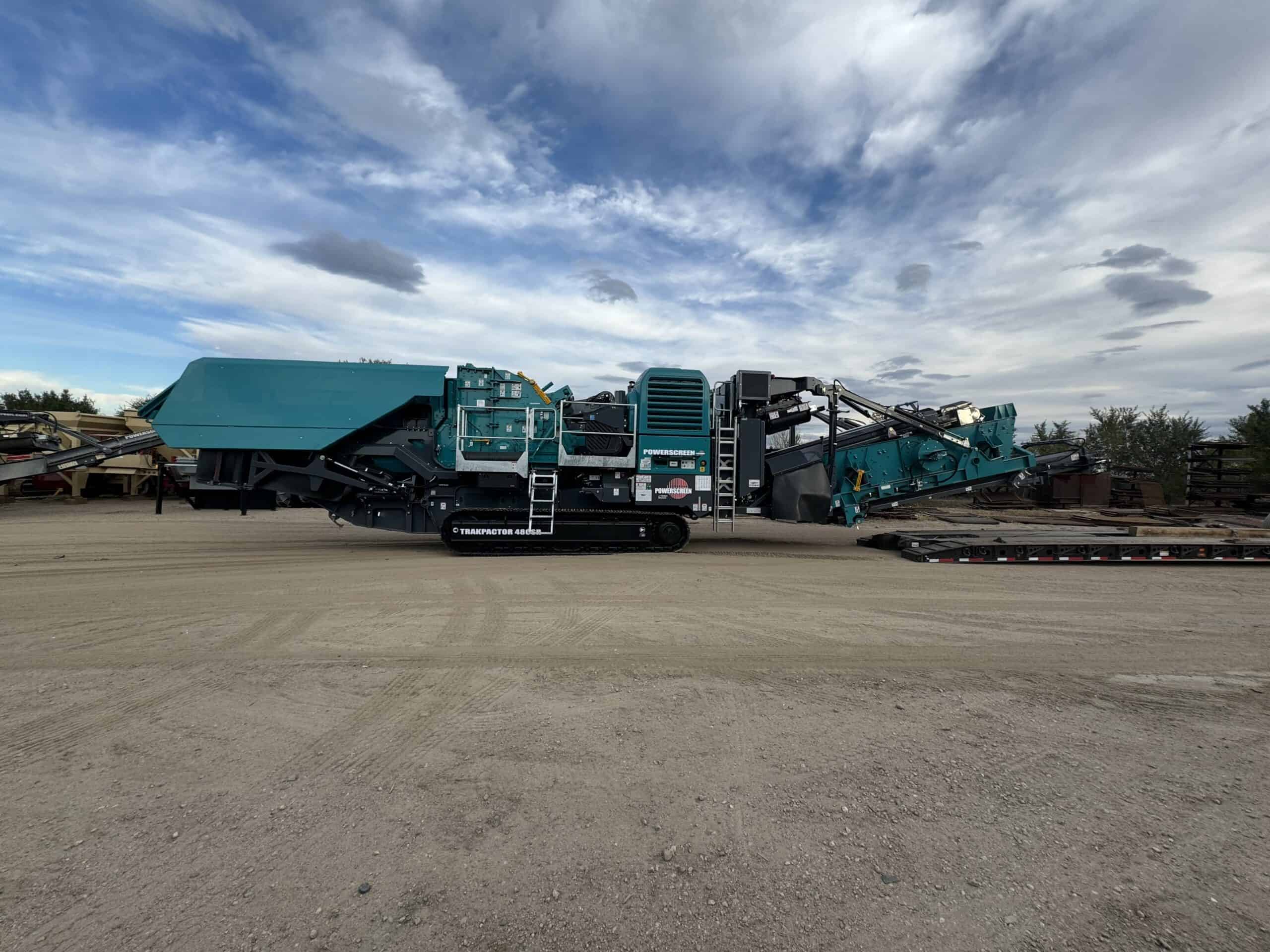 A Powerscreen Trakpactor 480SR mobile crusher positioned beside a lowboy RGN trailer, prepared for loading and heavy haul transport at an industrial equipment yard under partly cloudy skies.