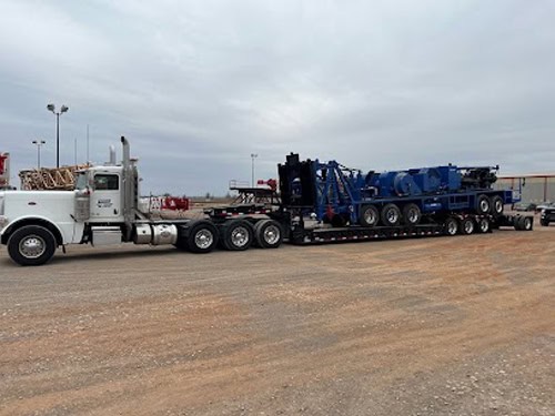 White truck hauling blue workover rig on multi-axle RGN trailer.